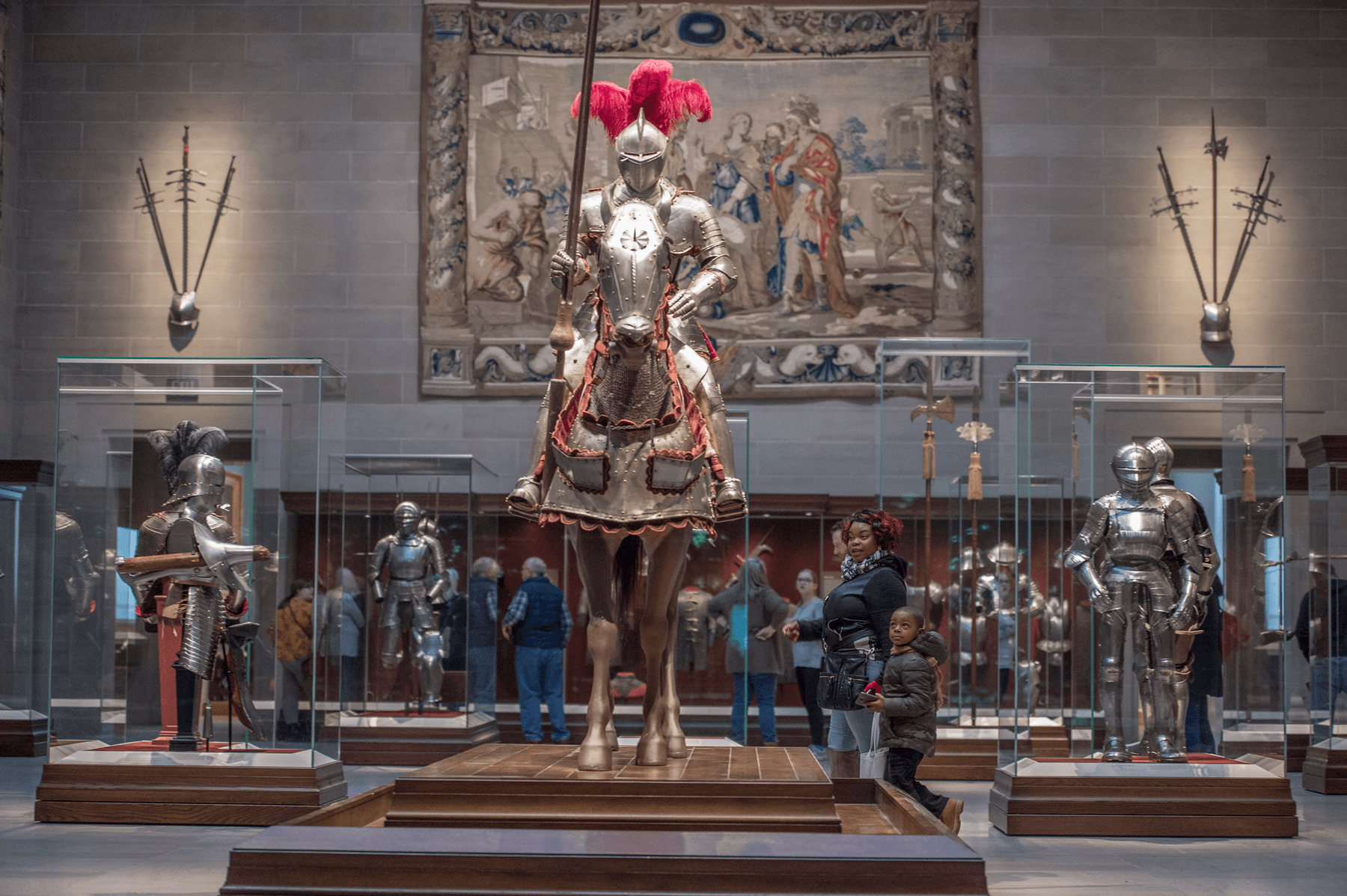 An adult and child standing near a life size mannequin of a horse and rider in armor, on view in a gallery with stone floors and walls, tapestries and armor and weapons displayed.