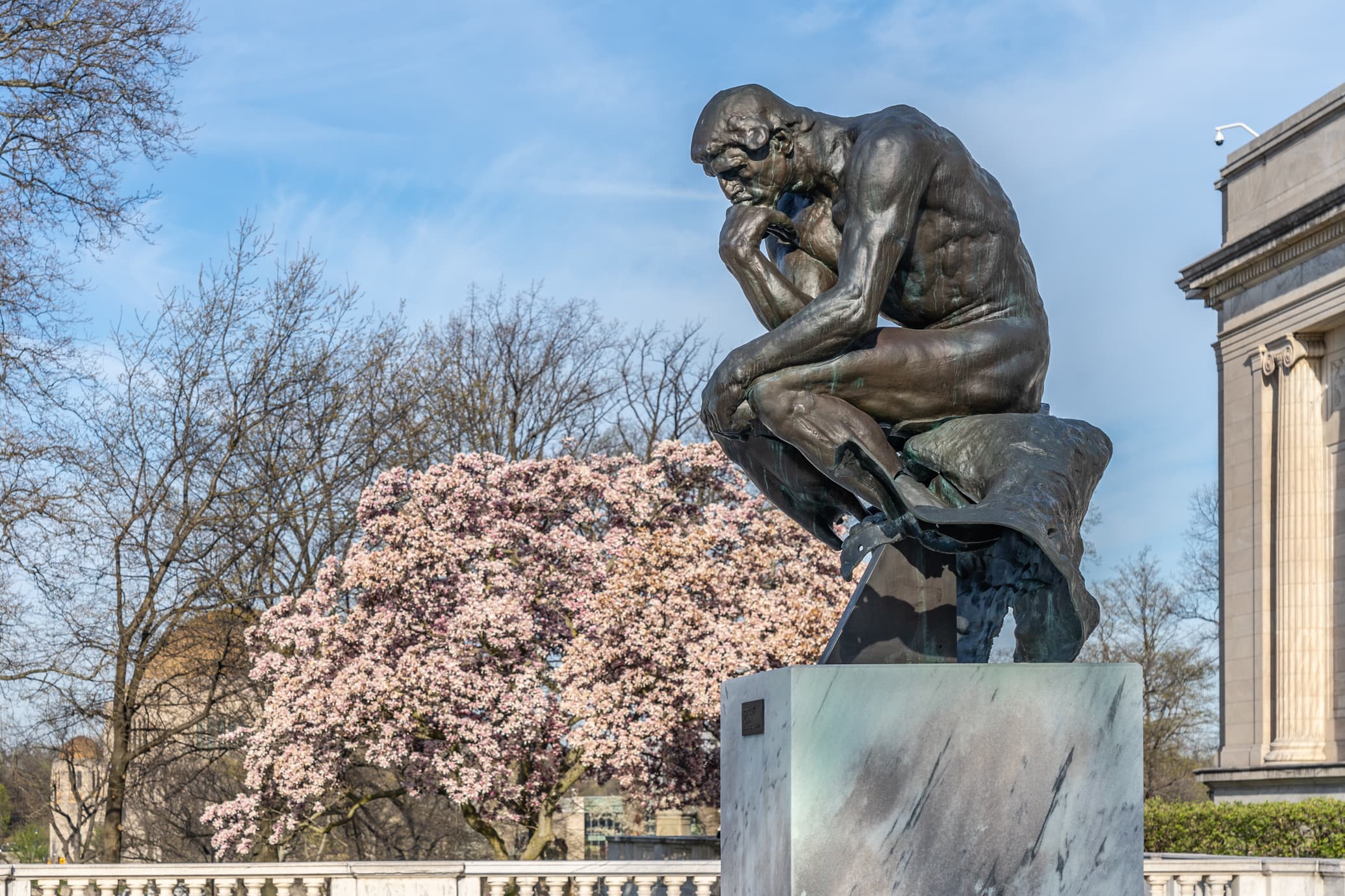 bronze sculpture of seated figure, chin resting on hand, installed outdoors on marble terrace