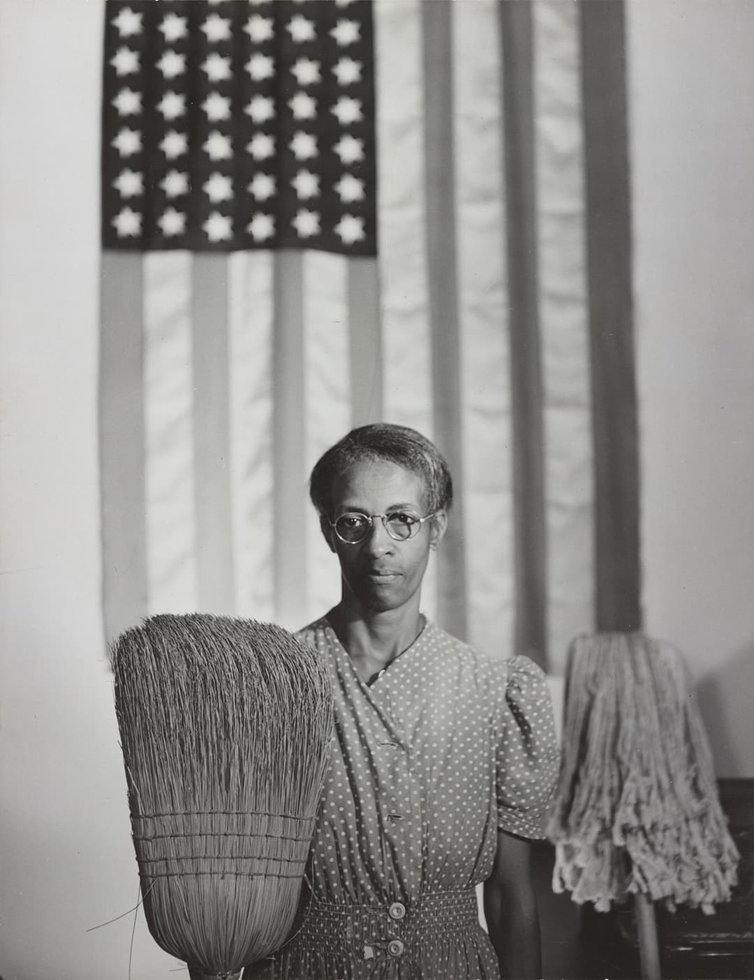 A woman holding a broom, with the straw up, in one hand and a mop, with the business end up, in the other hand. She is posed in front of an American flag.