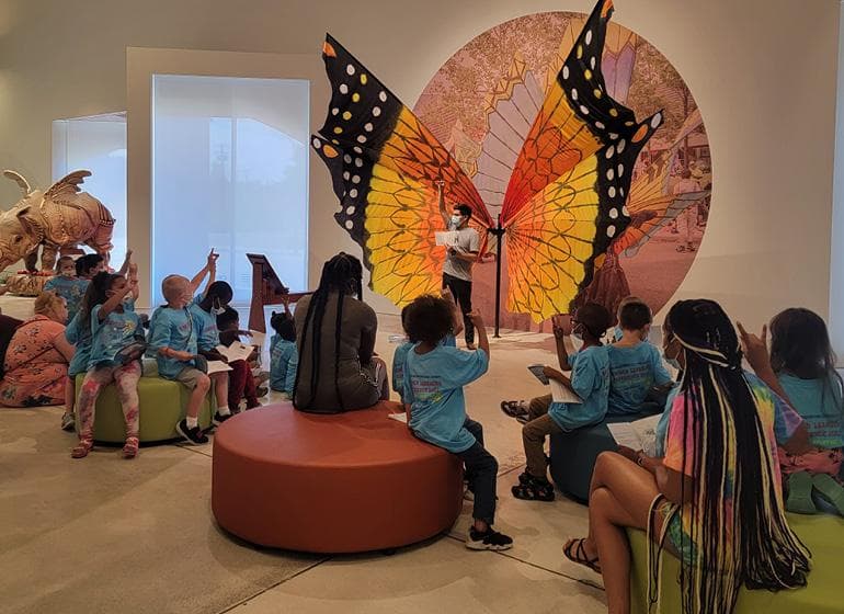 Students listen to educator, standing in front of larger-than-life mounted butterfly wings in gallery