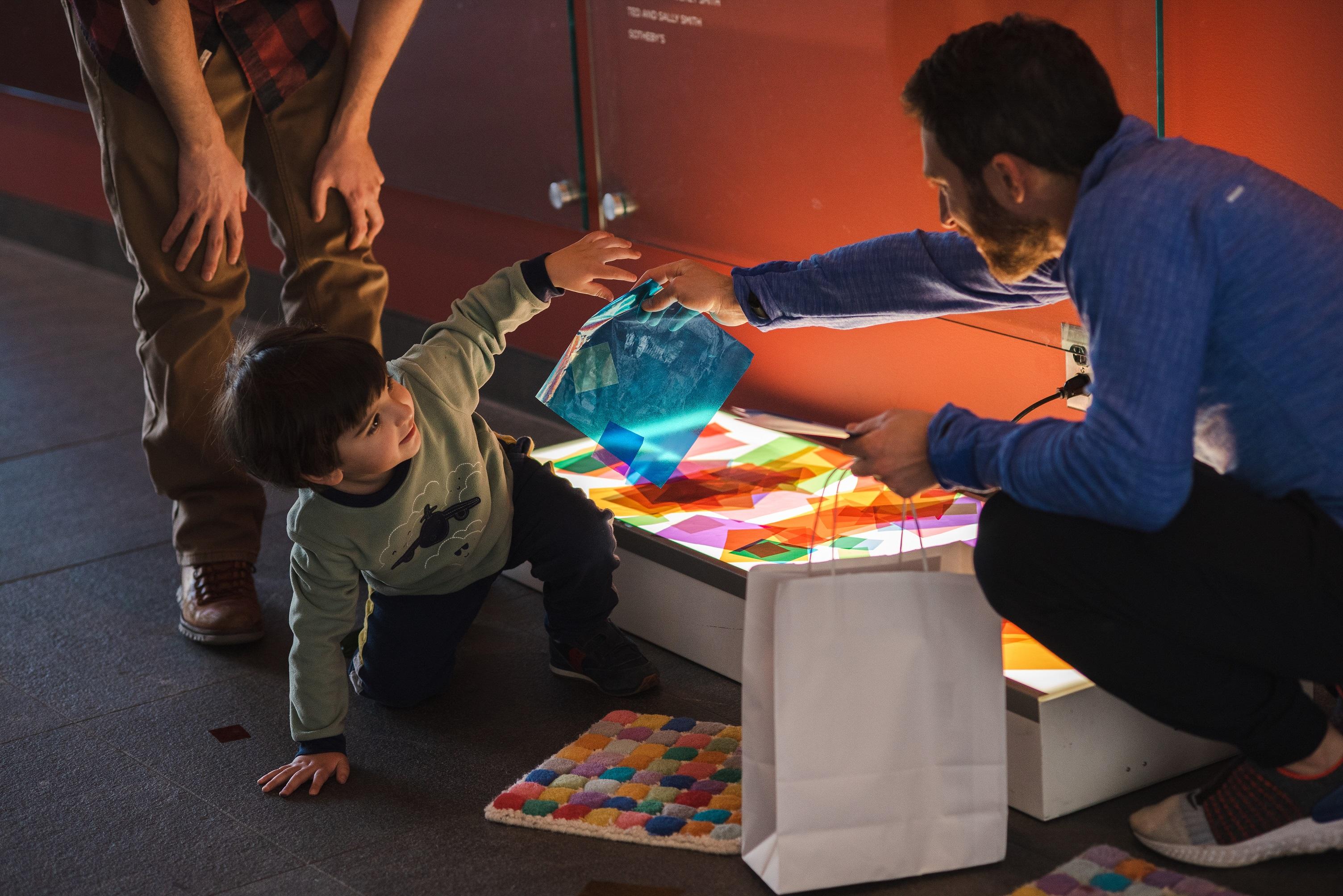 Child playing with colored cellophane on a light box and handing a piece of cellophane to an adult with them.