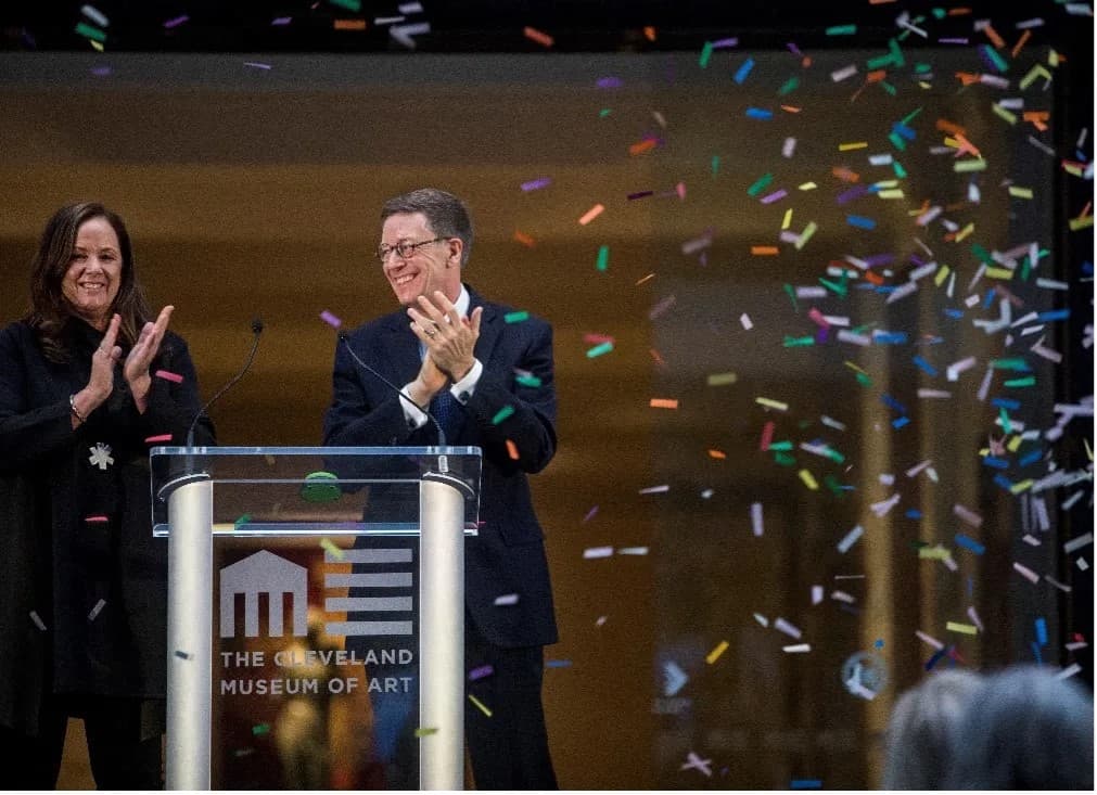 A man with light skin tone wearing a black suit and glasses clapping and standing next to the right of a woman with light skin tone and brown hair also clapping as there is colorful confetti raining down.