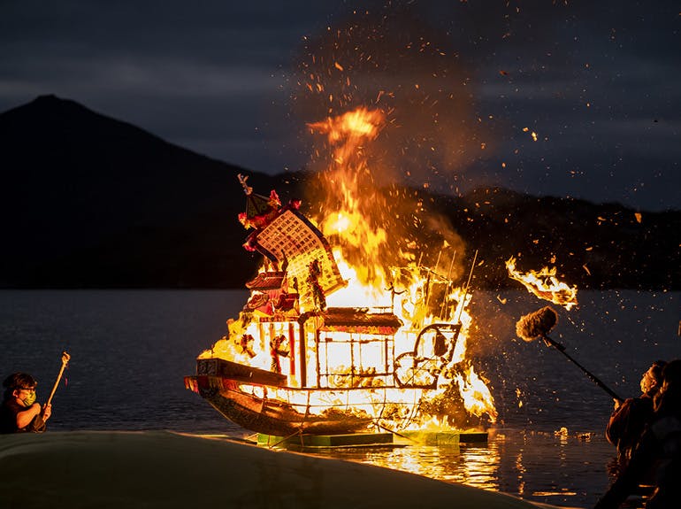 An image of a boat burning at night in a mountain lake, with two recording figures on either side