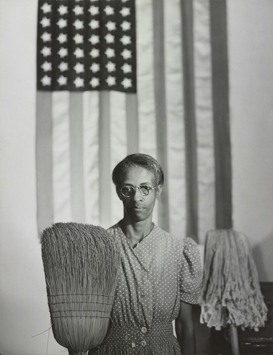 A woman holding a broom, with the straw up, in one hand and a mop, with the business end up, in the other hand.  She is posed in front of an American flag.  