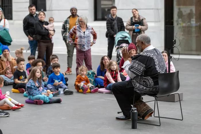 A storyteller sitting on stool in front of children in the atrium. The storyteller is playing a drum.