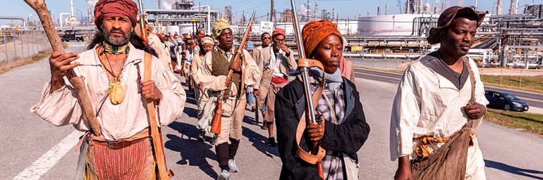 procession of people dressed historical clothing, marching through an industrial landscape.